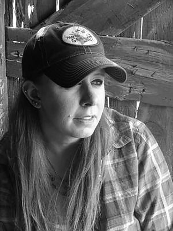 Woman in plaid shirt and cap gazes off-camera, set against a rustic wooden structure.