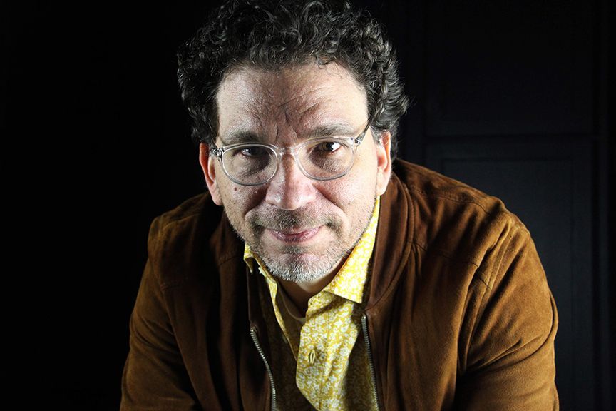 A man with curly hair and glasses is wearing a brown jacket and a yellow shirt.
