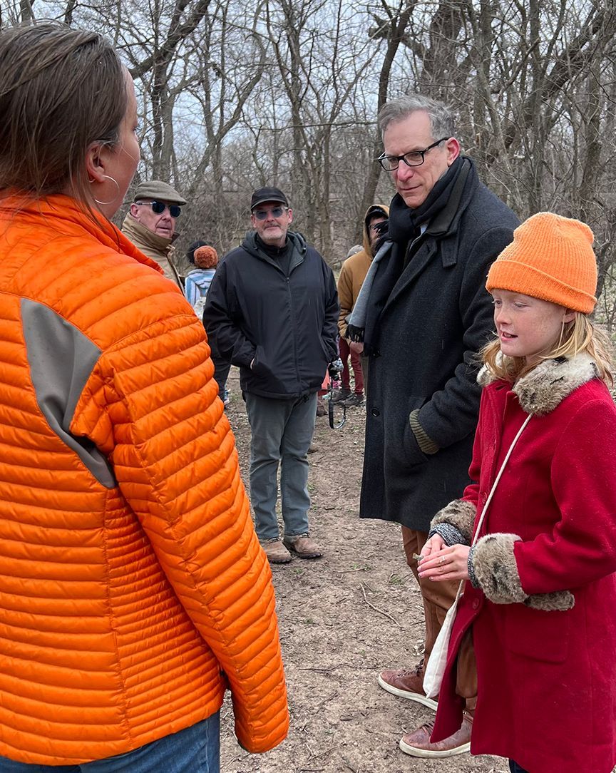 People talking outdoors, child in red coat, woman in orange jacket, trees.