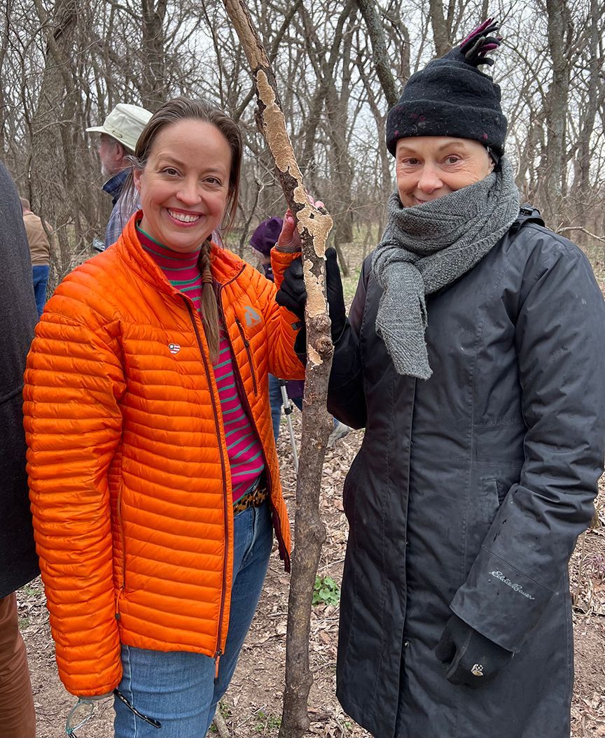 Two women smile, pose with a small tree in a wooded area. One wears orange jacket, the other a black coat and hat.