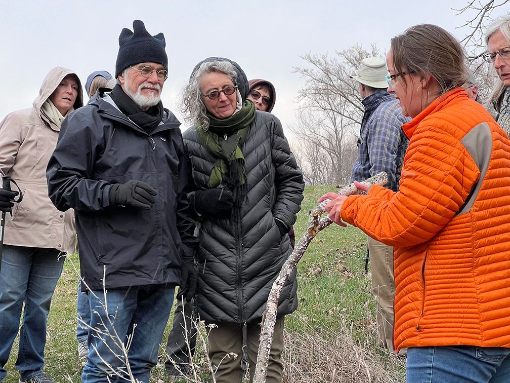 Group of people on a nature walk, listening to a person in an orange jacket. Outdoors, cloudy day.