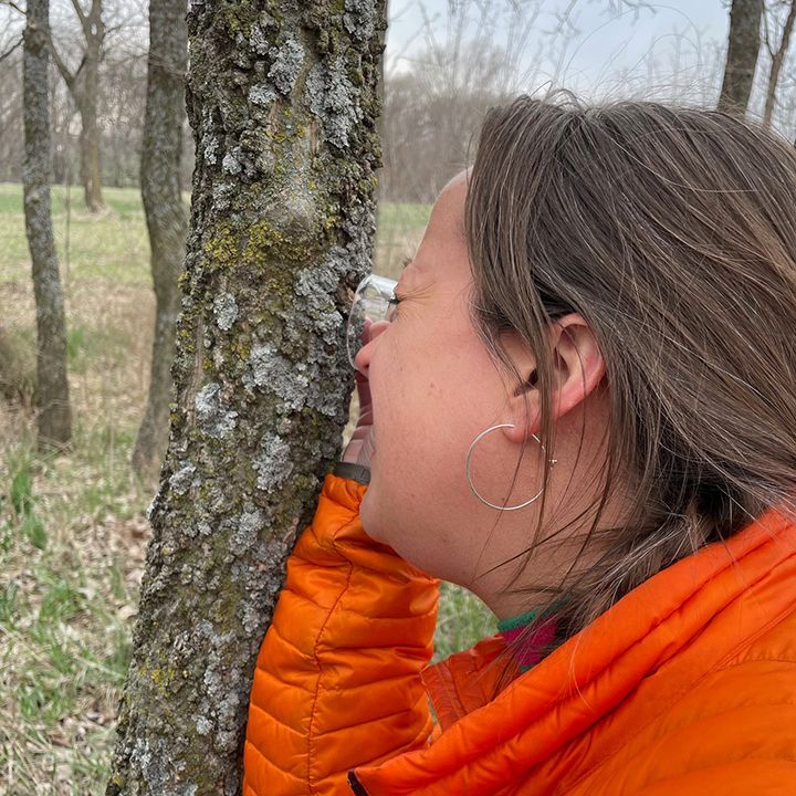 Woman with orange jacket looks closely at a tree trunk outside.