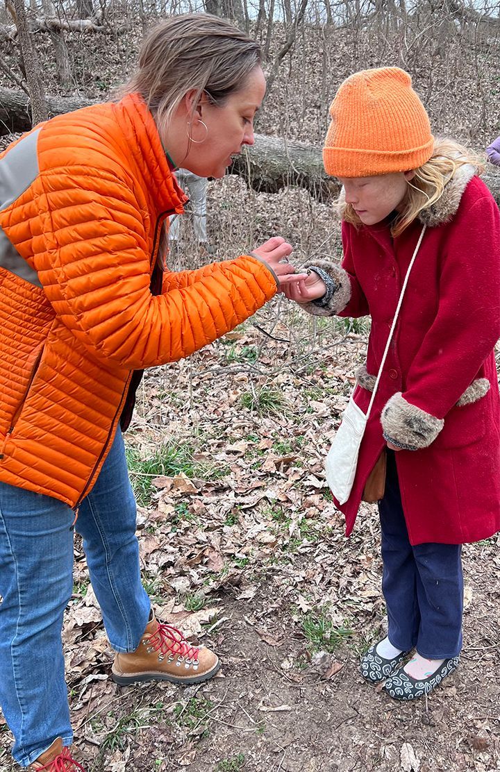Woman in orange jacket shows a tiny object to a girl in a red coat and orange hat, outdoors on a trail.
