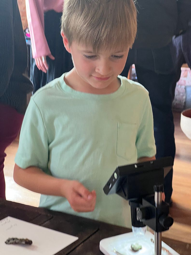 Boy in light blue shirt looks at small object through a microscope at a wooden table.