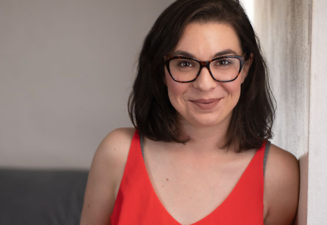 Woman with glasses, red top, smiles at the camera, leaning against a wall indoors.