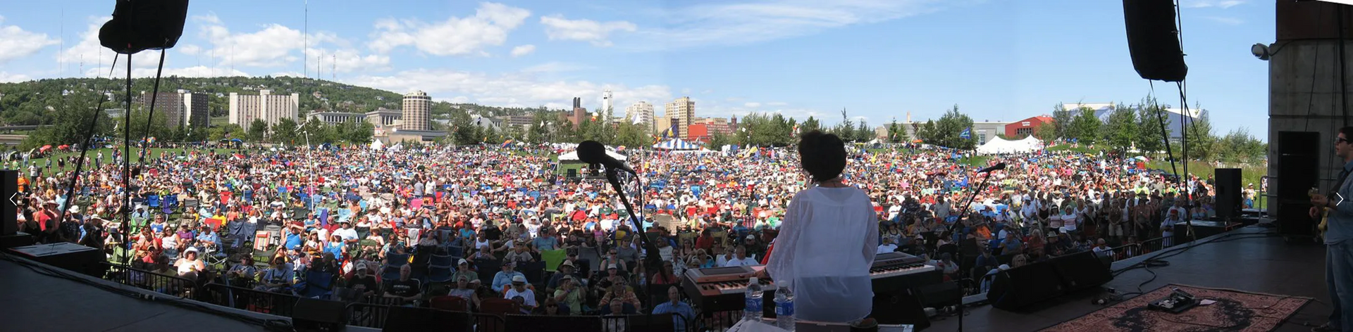 A performer faces a large crowd at an outdoor concert venue. Sunny day with city buildings in the background.