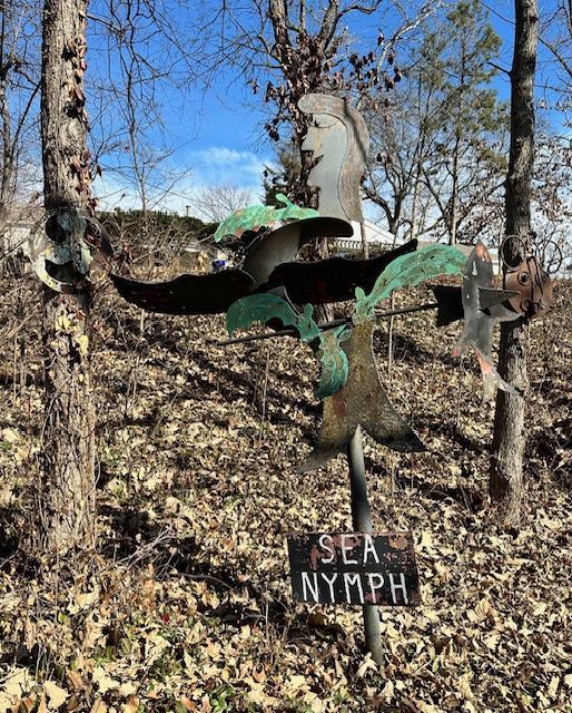 A metal sculpture of a sea nymph in a wooded area with a sign.