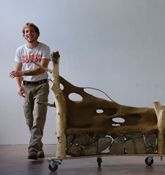 Man smiling next to a wheeled, wood-and-stone bench sculpture. Beige wall background.