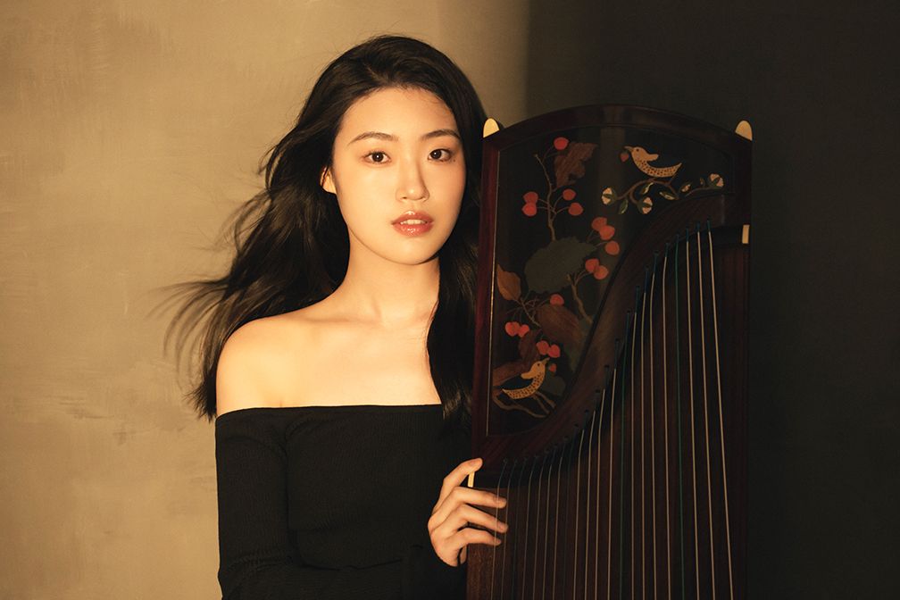 Woman with dark hair in black top, holding a decorated guzheng against a neutral background.