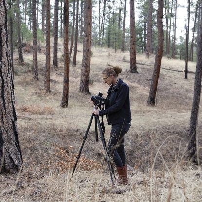 Woman with camera on tripod in a forest, focused on taking a picture.