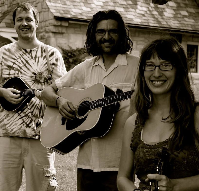 Three musicians with instruments: mandolin, guitar, and violin, smiling outside a stone building.