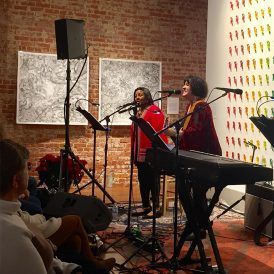 Two women singing, performing in front of a seated audience. Brick wall, abstract art, and colorful bird art in the background.