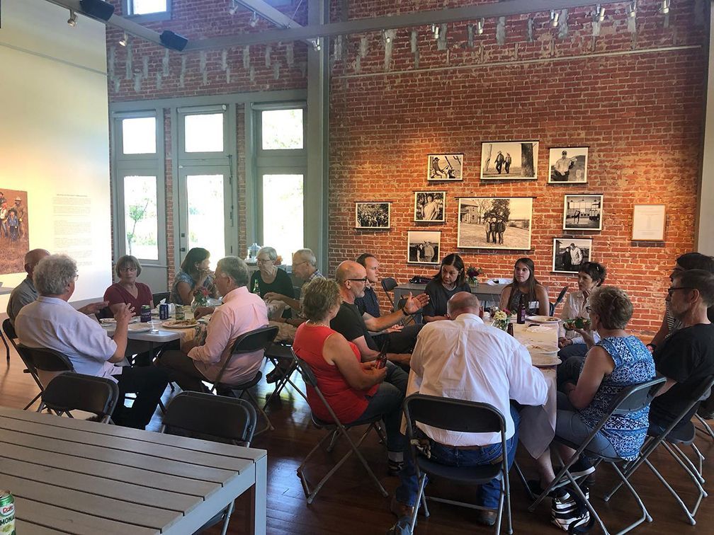 Group of people sitting at round tables in a gallery, conversing. Red brick wall with framed photos in background.