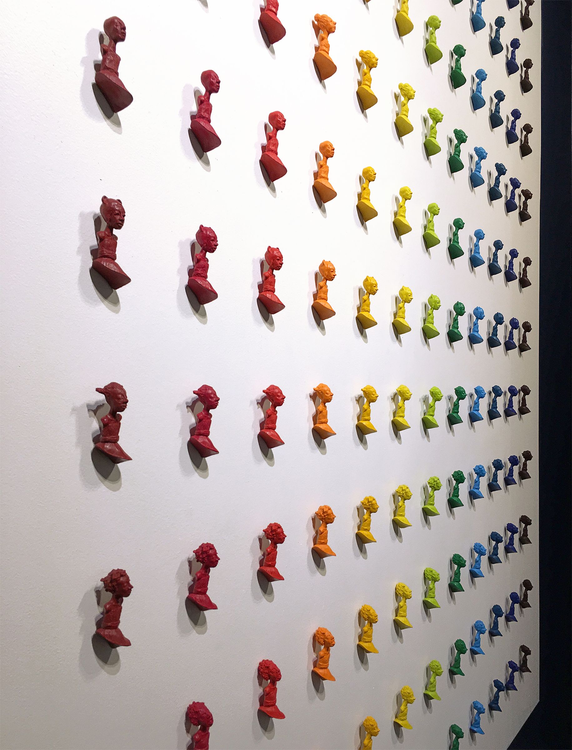 Rows of colorful, abstract sculptures mounted on a white wall; red to blue color gradient.
