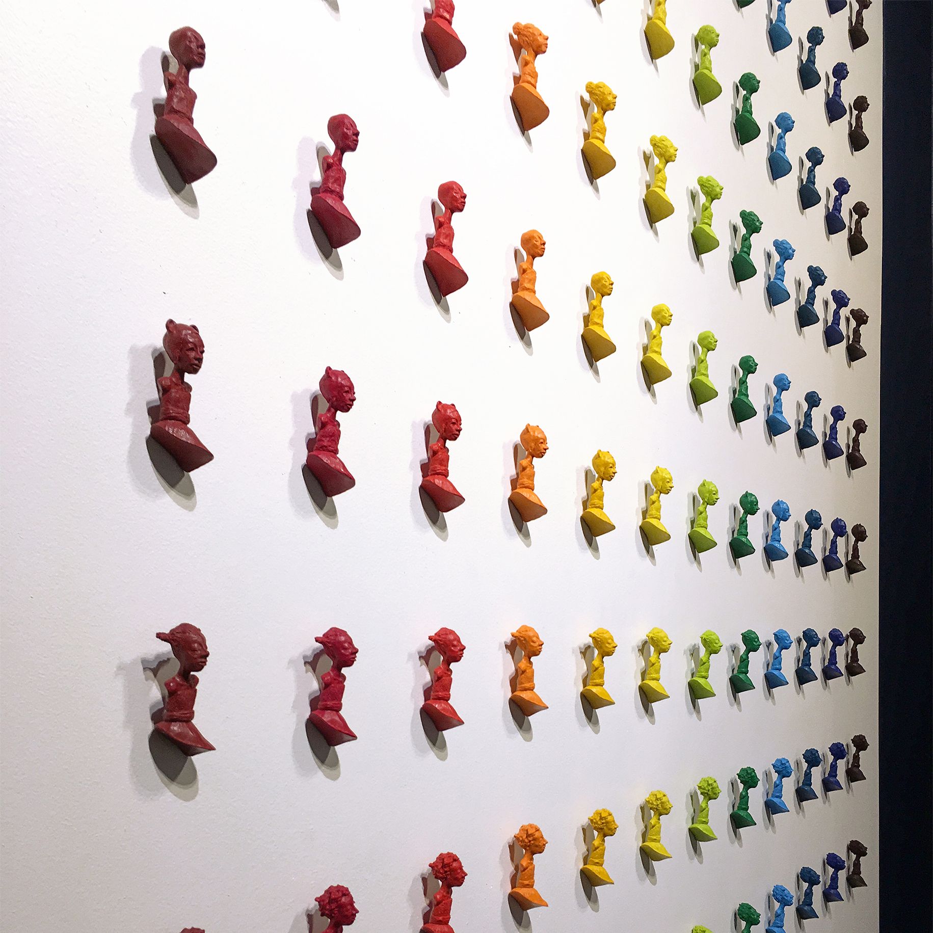 Rows of colorful, stylized figures arranged on a white wall in a rainbow pattern.