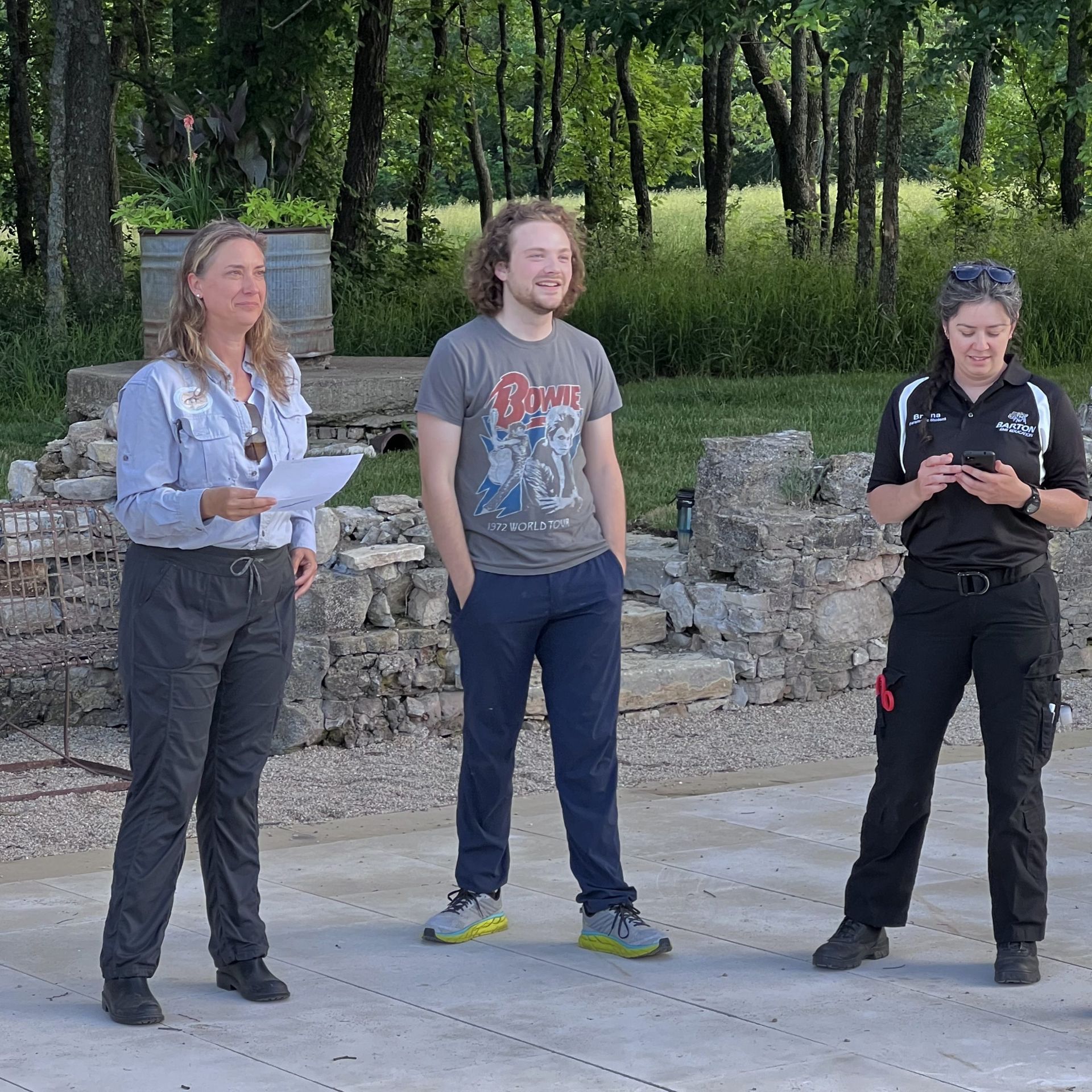 Three people stand outdoors near stone ruins. One holds papers, one smiles, and one looks at a phone.