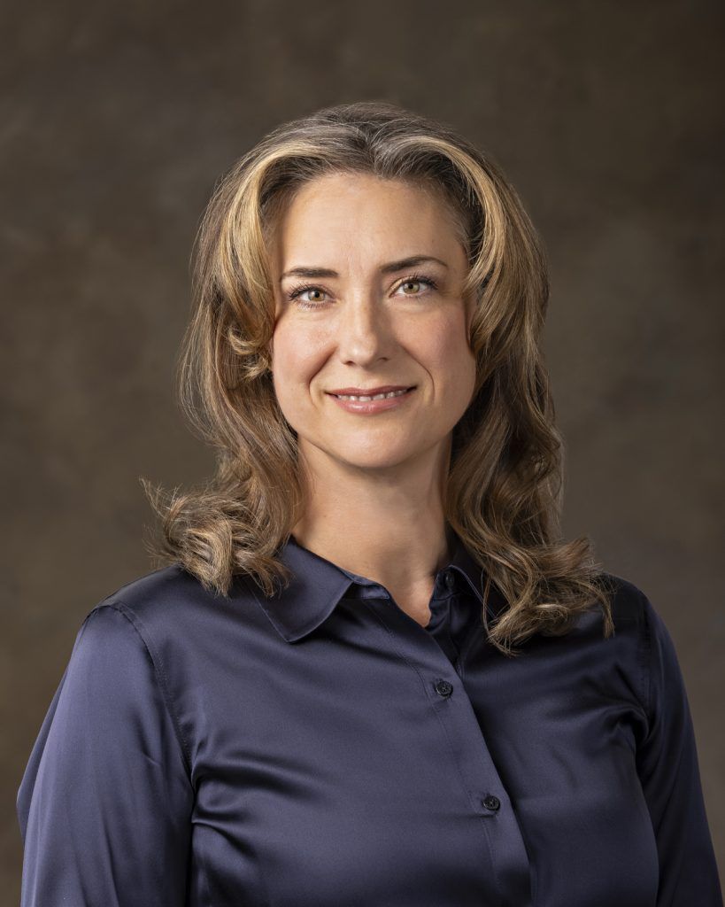Woman with brown hair in a blue blouse, smiling at the camera, against a brown background.