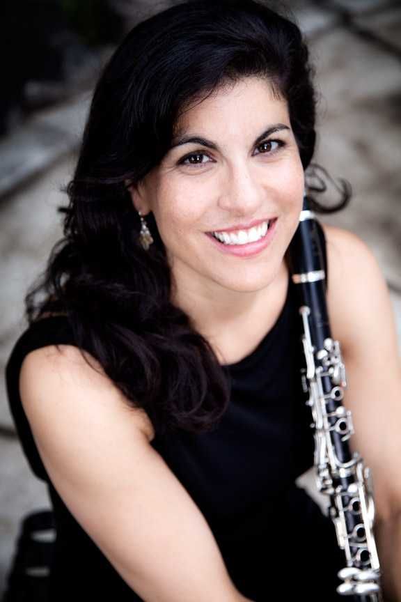 Woman with dark hair smiles, holding a clarinet. She wears a black dress and sits outdoors.