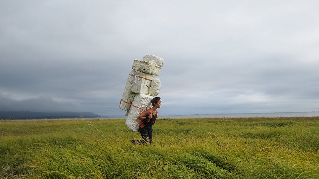 Man carrying a tall stack of white packages across a grassy field under a cloudy sky.