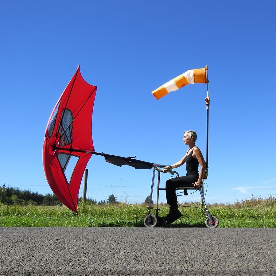 Person using a walker with a sail attached, rolling down a road. A windsock points toward the wind.