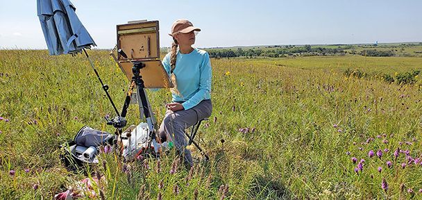 A woman paints outdoors in a field of green grass with purple flowers, on a sunny day.