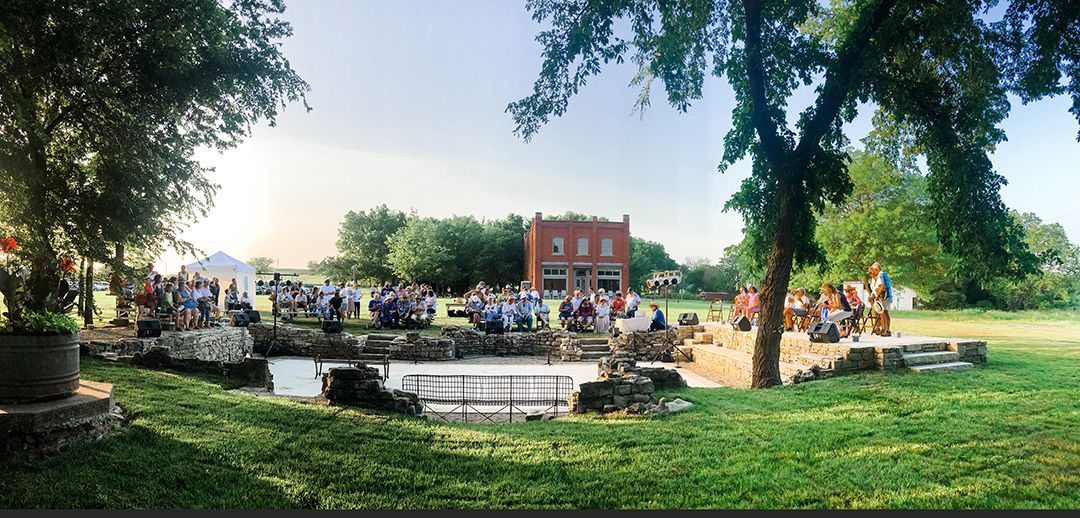 People gathered outdoors near a small pond and red brick building. Trees frame the event.