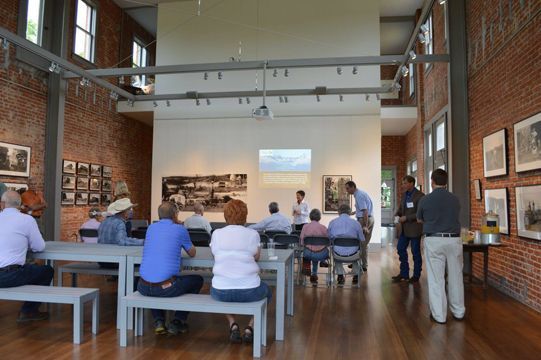 People seated at tables in a brick-walled room, a presentation displayed on a screen.