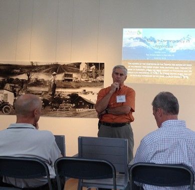 Man giving presentation with two seated listeners. Room with photo and projection screen visible.