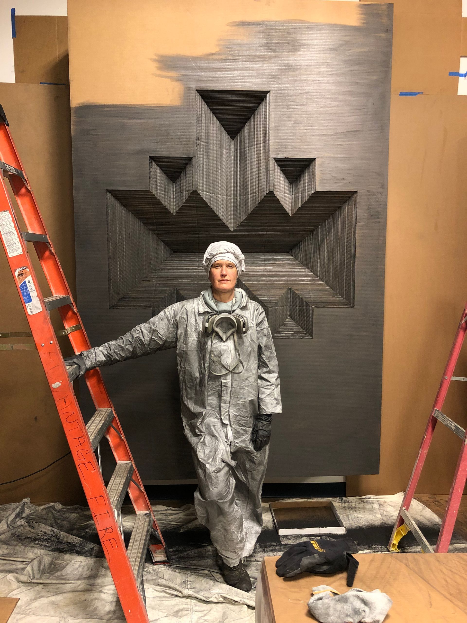 Person in protective suit stands before large geometric artwork, ladders on either side.