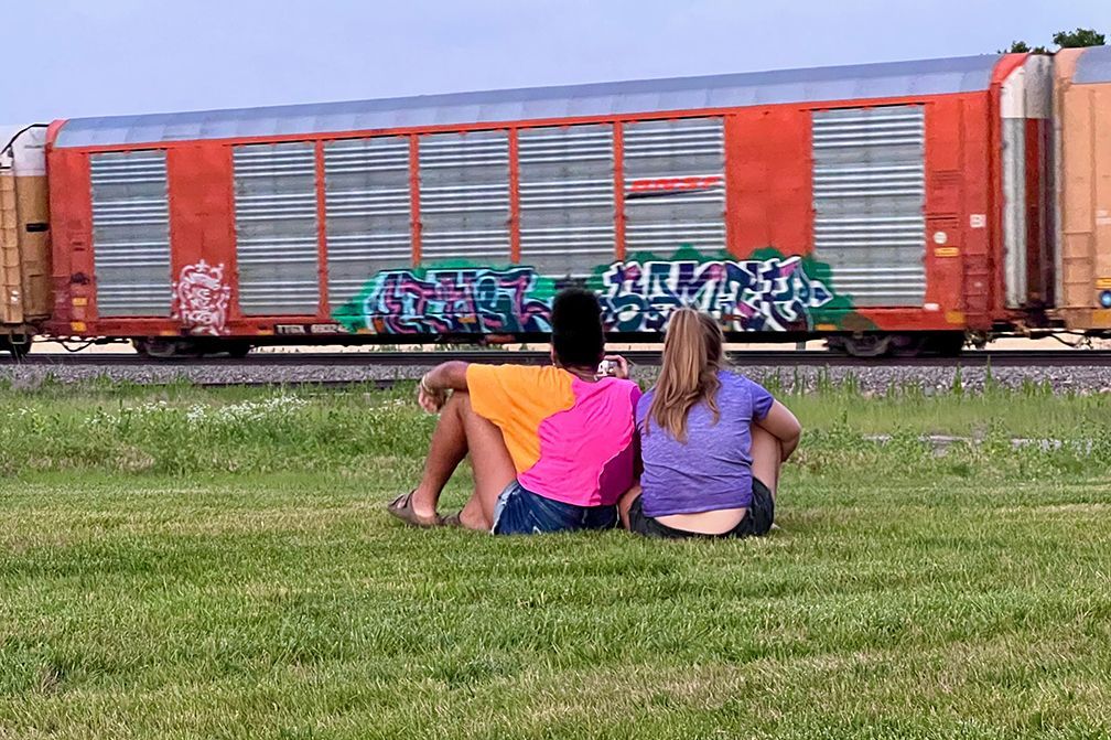 Two people sit on grass, facing away from the camera, watching a passing freight train.