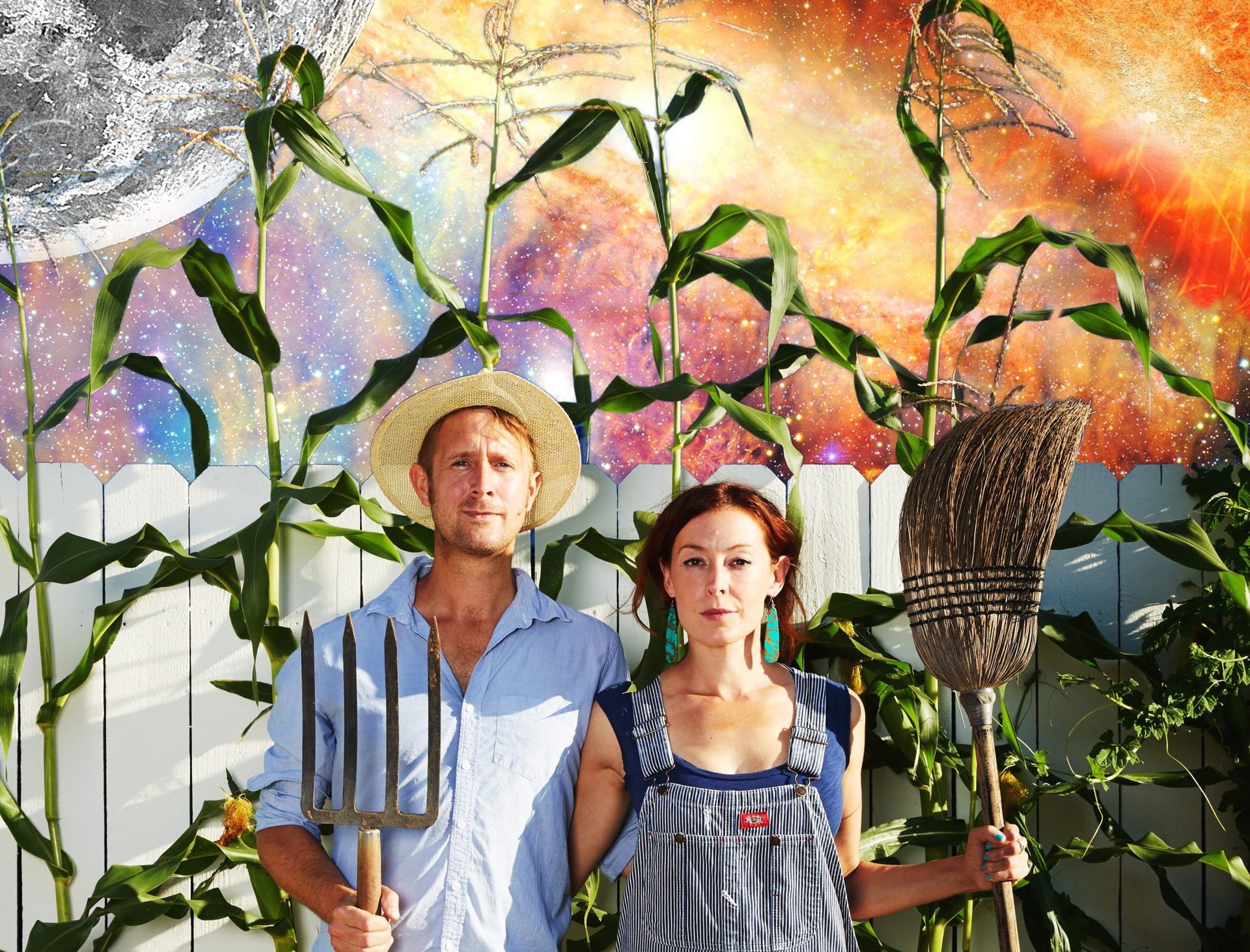 Couple in front of mural holding farm tools; corn stalks frame them. Colorful celestial scene backdrop.