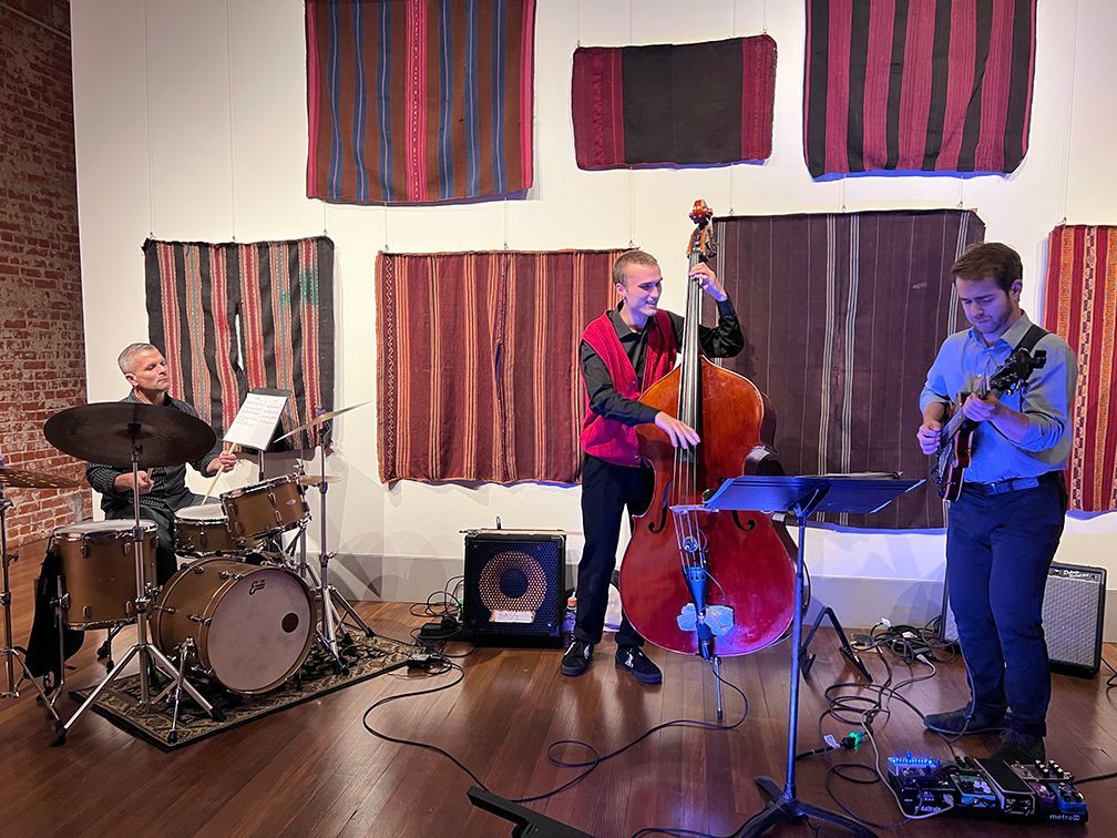 Jazz trio performing on a stage: drummer, bassist, and guitarist, with hanging textile art in the background.