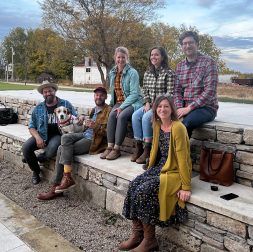 Group of six people and a dog sitting on a stone wall. Autumn day, mostly neutral colors.