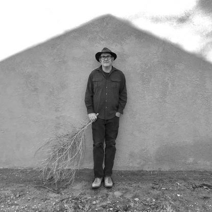 Man in hat, glasses, and casual clothes standing against a wall, holding a leafy branch.