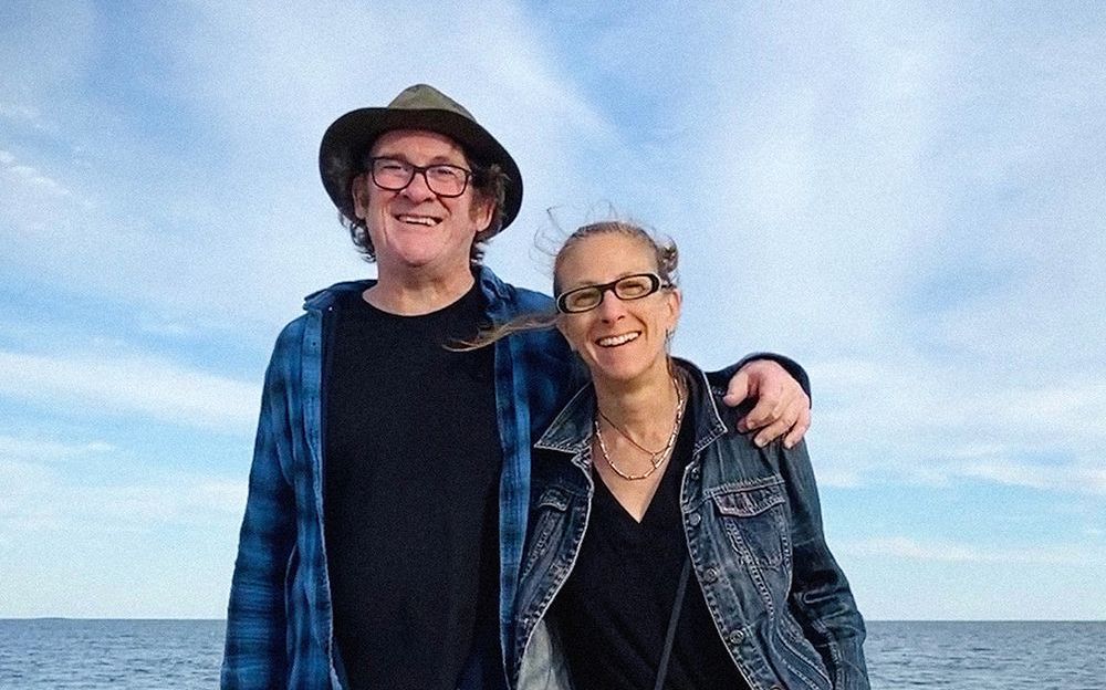 Man in hat and woman with glasses smiling, ocean background.