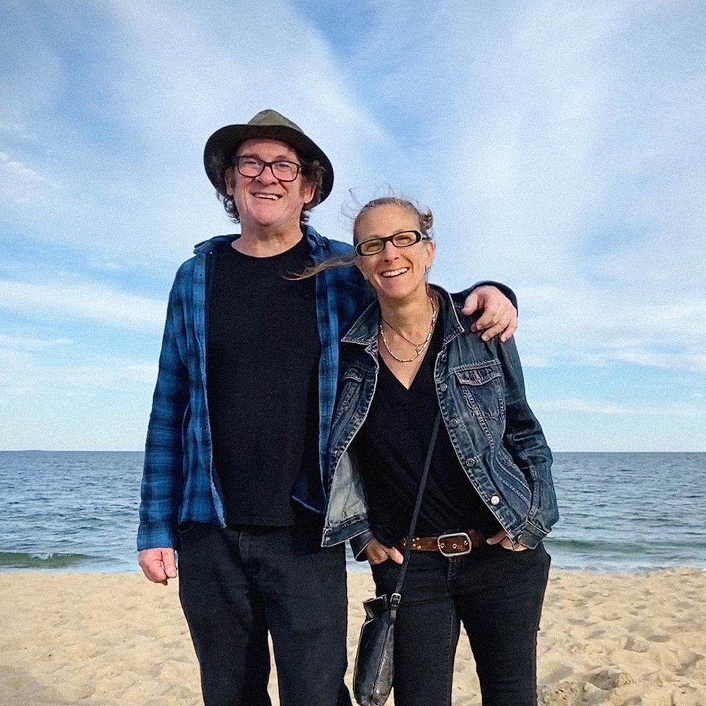 A man and woman stand on a beach, smiling. The man has a hat and the woman is wearing glasses. Ocean in background.