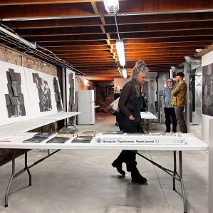 An art exhibition in a basement. A woman looks at work on a table, two others stand by a wall.