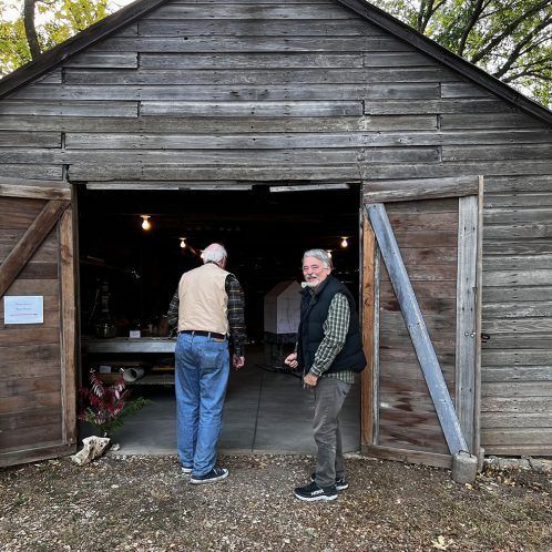 Two men stand in the doorway of a weathered wooden shed. One man is entering, the other is exiting.