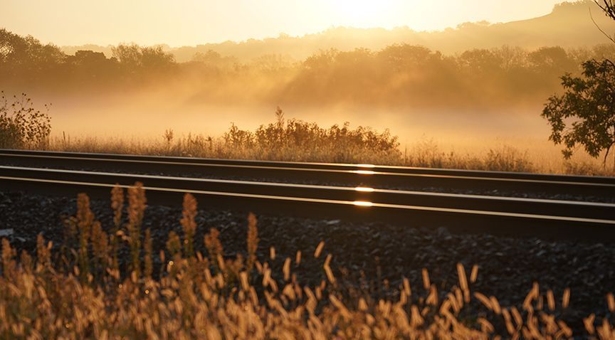 Train tracks through golden fields with a foggy, sunlit landscape.