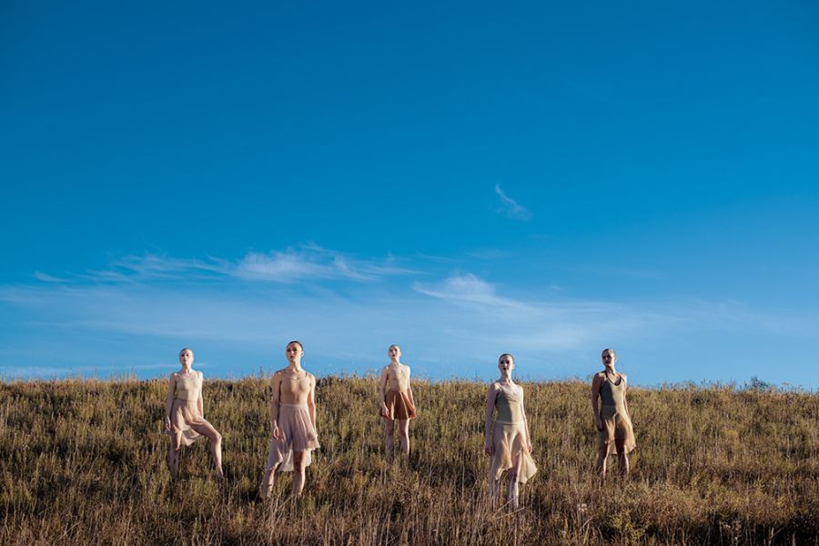 Five figures in flowing dresses on a grassy hill under a bright blue sky.