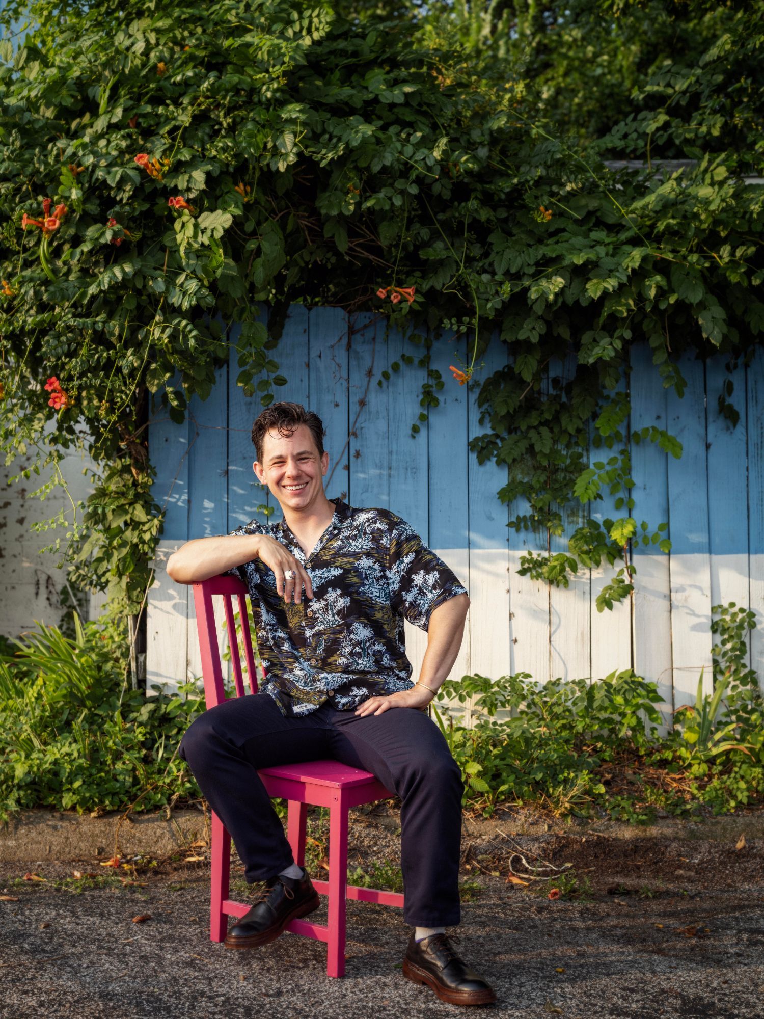 Man in floral shirt seated on pink chair, smiling, against blue and white wall with greenery.