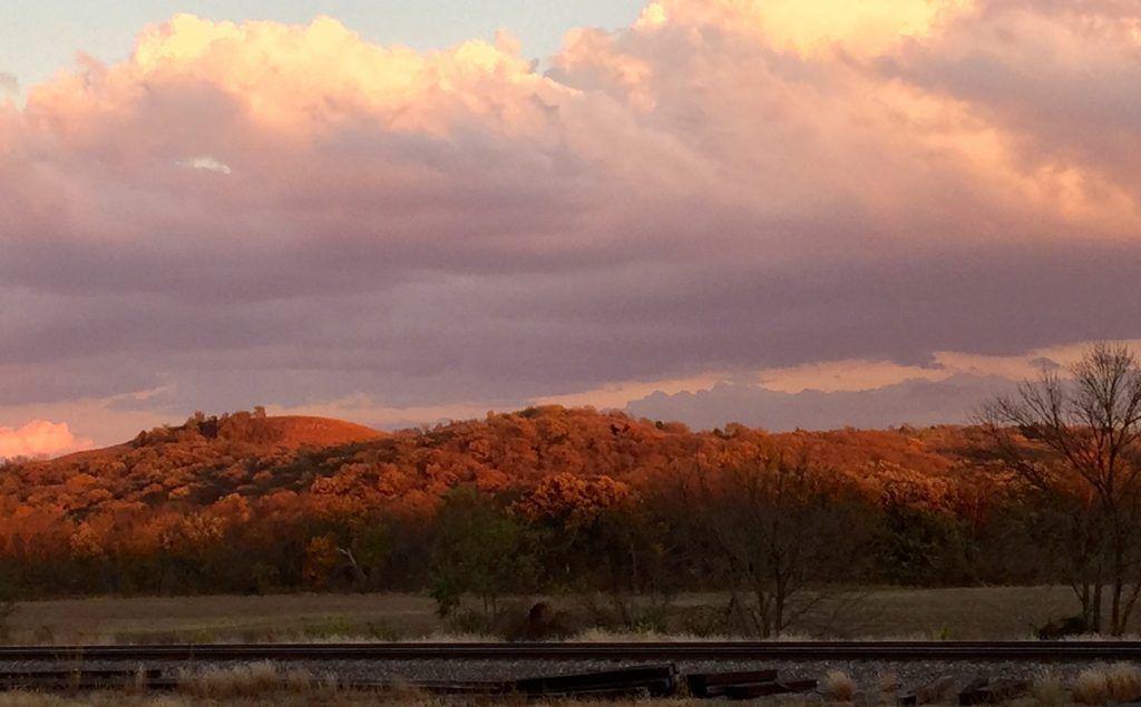 Sunset over a treed hill with railway tracks in the foreground. Clouds are pink and orange.