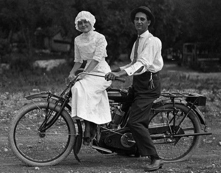 Couple on a vintage motorcycle; woman in white dress, man in shirt and tie; outdoors.