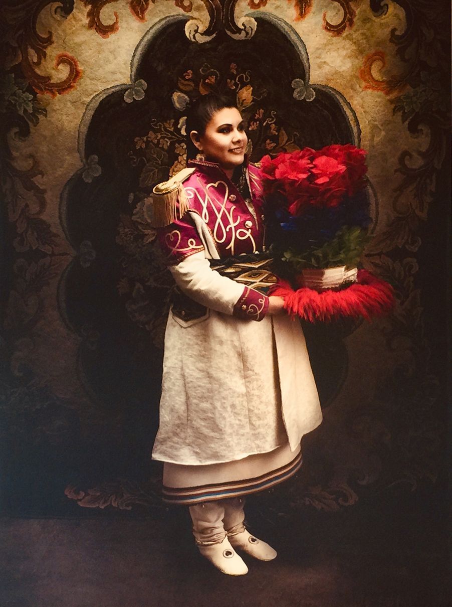 Woman in traditional attire holds a large bouquet of red flowers, smiling against an ornate backdrop.