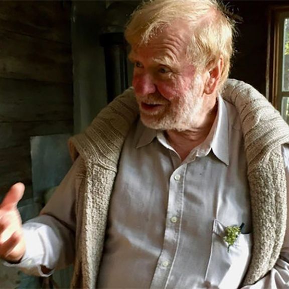 Elderly man with white beard and blond hair, gesturing, wearing a sweater, standing near a window.