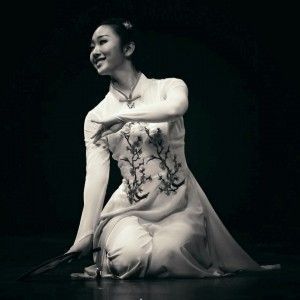 A woman in a white dress is kneeling down on the floor holding a fan.