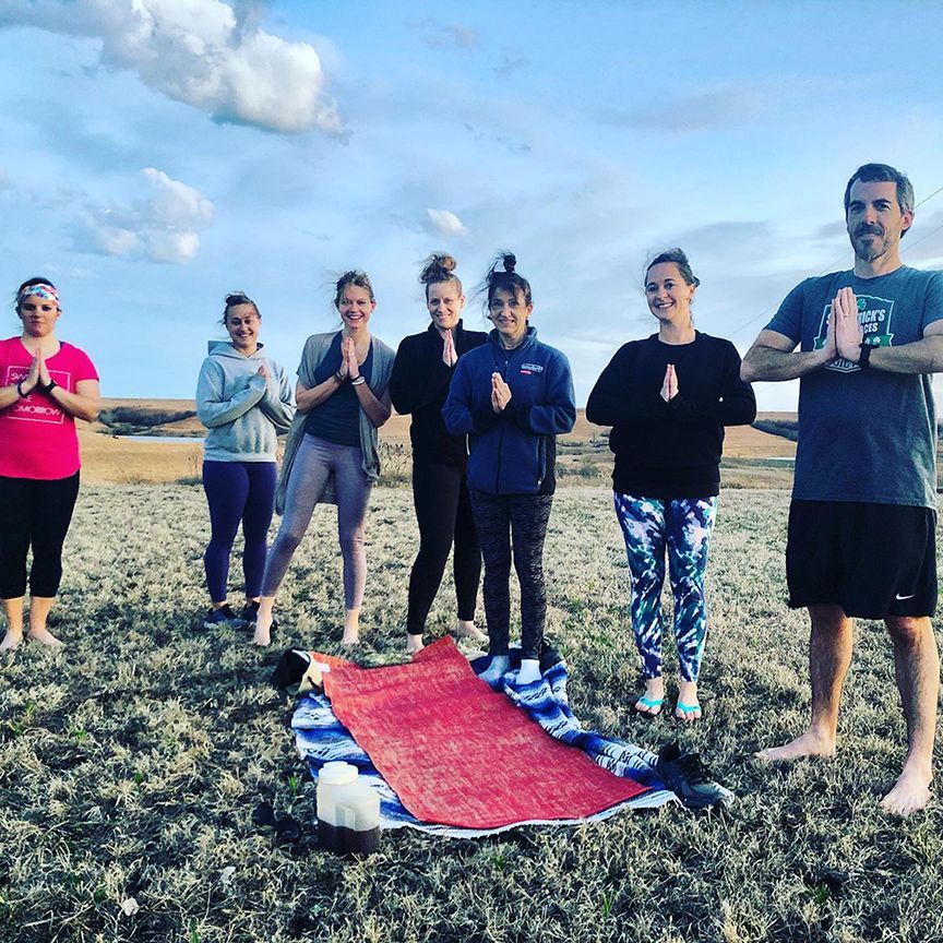 Group of people in outdoor yoga pose, hands in prayer, on a field. Blue sky, neutral tones.