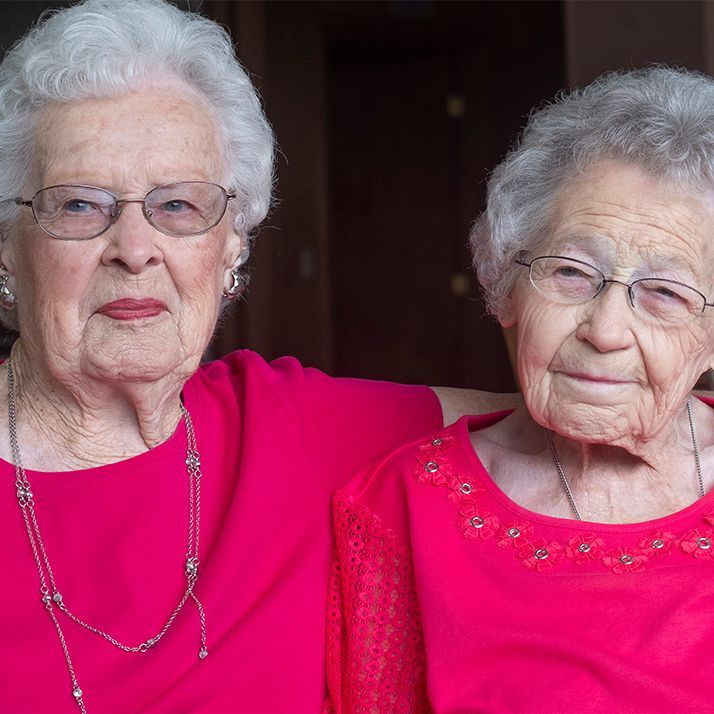 Two elderly women, wearing glasses and red tops, pose together indoors.