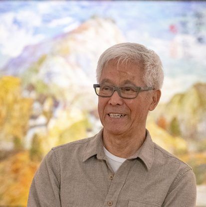 Smiling elderly Asian man with glasses, tan shirt, in front of a colorful mountain painting.