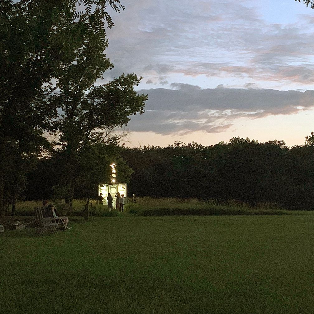 Dusk scene: illuminated glass structure in a field with people, shadowed trees, and a cloudy sky.